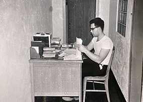 A young man in profile sits at his desk, shuffling papers.  He’s wearing a white tee shirt, black pants and glasses.